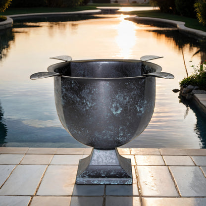 A metal cigar ashtray with an antique distressed finish, featuring four stirrups and a deep bowl design, placed on a stone surface with a body of water and sunset in the background.