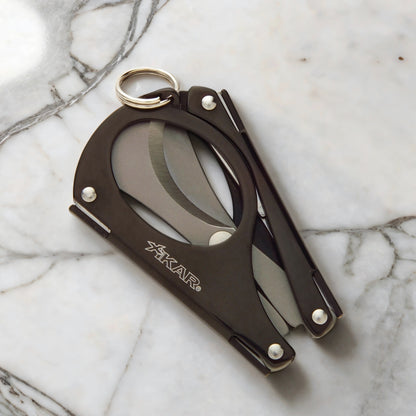 A black, foldable multi-tool cigar scissors with stainless steel blades, featuring the XIKAR brand name on the side.