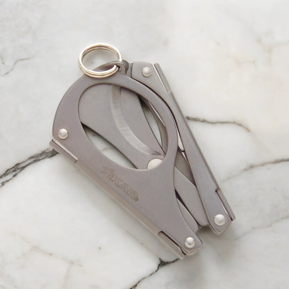 A foldable, stainless steel multi-tool cigar scissors with a ring on the top for easy carrying, displayed on a marble surface.